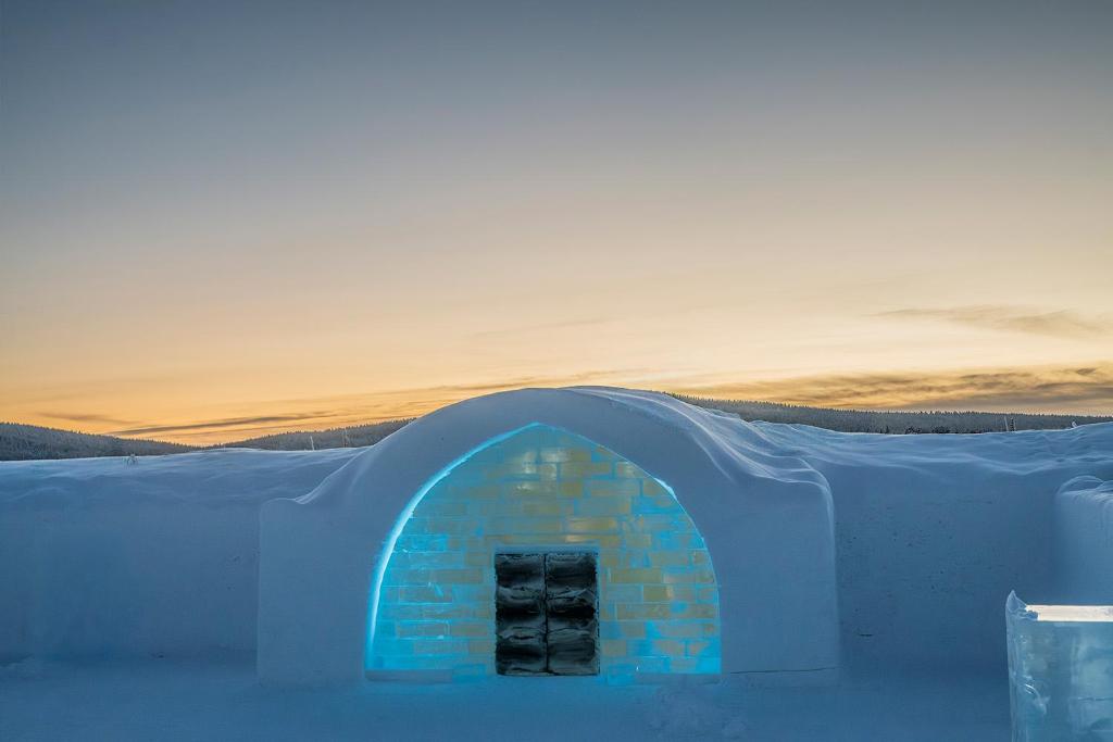 A low timber cabin in snow, lit from within, at the end of the afternoon.